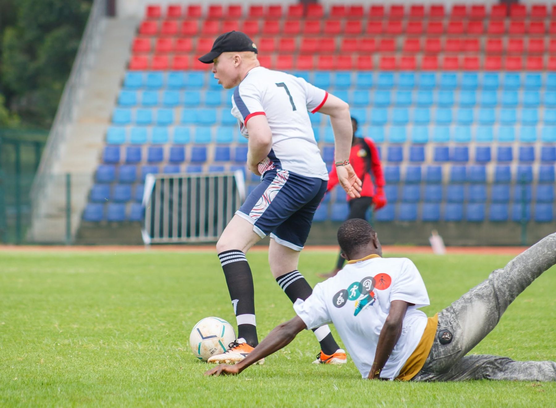 A Black Albinism footballer in a white number seven shirt and cap takes the ball past a sliding defender on a grass pitch.