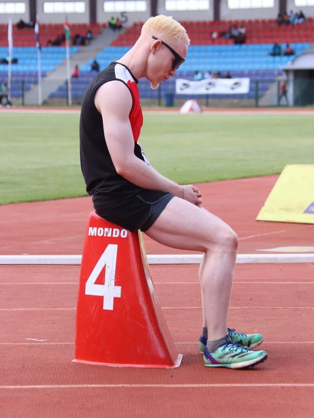 A young man with albinism rests on a starting block at the edge of a red running track after a race.